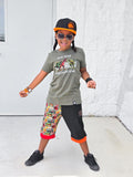 Child wearing a green t-shirt with graphics, colorful shorts, and a black with orange accents Jurassic Snapback.