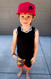 Child wearing a red Jurassic Snapback and black sleeveless shirt with colorful shorts standing on a concrete surface.