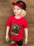 Child wearing a red t-shirt with a dinosaur graphic and a black and red Jurassic Snapback against a brick wall.