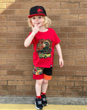 Child wearing a red t-shirt with a dinosaur graphic, a red Jurassic Snapback and black shorts against a brick wall.