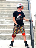 Child wearing a black t-shirt with a graphic design, colorful shorts, and a Jurassic Snapback on a set of stairs.
