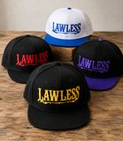 Four snapback caps arranged on a wooden surface against a neutral wall. One black cap in front has yellow 'LAWLESS' embroidery, one black cap at left has red 'LAWLESS' embroidery, one black cap at right has purple 'LAWLESS' embroidery, and one white cap with blue brim and blue 'LAWLESS' embroidery stands behind on a small wooden block.