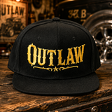 A black flat-brim cap with 'OUTLAW' embroidered in distressed gold lettering across the front, displayed on a rustic wooden surface. The background is a dimly lit, vintage-style garage or workshop with a hot rod and posters.