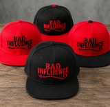 Four snapback caps arranged on a wooden surface against a gray wall, alternating red and black, each embroidered with the phrase 'BAD INFLUENCE' in red with a black arrow pointing left.