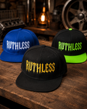 Three flat-brim snapback caps displayed on a rustic wooden table in a garage-like setting. The front cap is black with a yellow 'RUTHLESS' logo, the left cap is blue with a white 'RUTHLESS' logo, and the right cap is black with a neon green brim and a green 'RUTHLESS' logo.