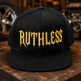A black snapback cap with the word 'RUTHLESS' embroidered in a gold gothic font across the front, displayed on a rustic wooden table with a blurred garage workshop background.