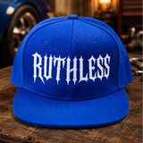 A bright blue snapback cap with the word 'RUTHLESS' embroidered in white gothic-style lettering across the front, displayed on a rustic wooden table with a blurred garage background.