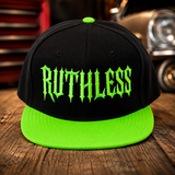A black flat-brim snapback cap with a bright neon green brim and matching green button, featuring the word 'RUTHLESS' in a green metal-style font on the front. The cap is photographed on a wooden surface with a blurred garage or workshop background and part of a car visible.