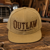 A tan baseball cap with 'OUTLAW' embroidered in dark brown across the front, displayed on a rustic wooden table inside a warm, wood-paneled garage or workshop.