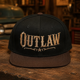 A black flat-brim snapback cap with 'OUTLAW' embroidered in a distressed gold font on the front, resting on a rustic wooden table with a vintage garage background.