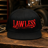 A black flat-brim baseball cap with 'LAWLESS' embroidered in a bold red gothic font on the front, displayed on a rustic wooden table with a blurred garage or workshop background.