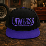 A black flat-brim snapback cap with a purple visor and purple embroidered 'LAWLESS' text on the front, displayed on a rustic wooden table with a garage workshop background.