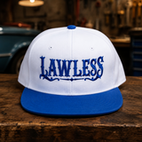 A white and blue flat-brim snapback cap with 'LAWLESS' embroidered in blue across the front, displayed on a rustic wooden table with a blurred garage background.