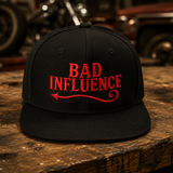 A black flat-brim baseball cap with red embroidered text reading 'BAD INFLUENCE' and a red arrow, displayed on a rustic wooden table with a blurred garage or workshop background.