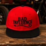 A red flat-brim snapback cap with black embroidery reading 'BAD INFLUENCE' and a black brim, displayed on a rustic wooden table with a blurred garage or workshop background.