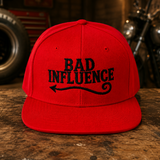 A bright red flat-brim baseball cap with black embroidered text reading 'BAD INFLUENCE' and a black arrow graphic, displayed on a rustic wooden table in a motorcycle-themed workshop setting.