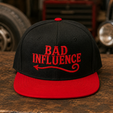 A black flat-brim cap with a red brim and red embroidered text reading 'BAD INFLUENCE' on the front, displayed on a rustic wooden table with a blurred garage or workshop background.