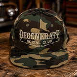 A camouflage baseball cap with 'DEGENERATE SOCIAL CLUB' embroidered on the front, resting on a rustic wooden table with a blurred workshop background.