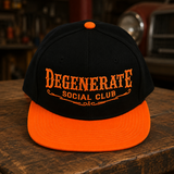 A black and bright orange snapback cap with 'DEGENERATE SOCIAL CLUB' embroidered in orange on the front, displayed on a rustic wooden table with a blurred workshop background.