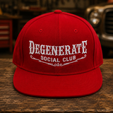 A red flat-brim baseball cap with white embroidered text reading 'DEGENERATE SOCIAL CLUB' on the front, displayed on a rustic wooden surface with a blurred garage or workshop background.
