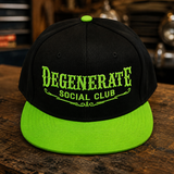 A black flat-brim snapback cap with a bright neon green visor, button, and embroidered text reading 'DEGENERATE SOCIAL CLUB,' displayed on a rustic wooden surface with a blurred garage or workshop background.