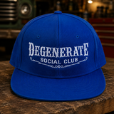 A royal blue flat-brim baseball cap with white embroidered text reading 'DEGENERATE SOCIAL CLUB' on the front, displayed on a rustic wooden table in a warm, blurred garage-like setting.