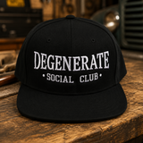 A black baseball cap with white embroidered text reading 'DEGENERATE SOCIAL CLUB' sits on a rustic wooden workbench with a blurred workshop background.