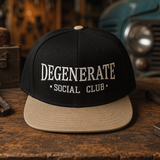 A black flat-brim snapback cap with beige brim and white embroidered text reading 'DEGENERATE SOCIAL CLUB', displayed on a rustic wooden table with a blurred workshop background.