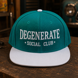 A teal and white snapback cap with 'DEGENERATE SOCIAL CLUB' embroidered on the front, displayed on a rustic wooden table in a vintage garage setting.