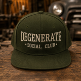 An olive green flat-brim baseball cap with 'DEGENERATE SOCIAL CLUB' embroidered in cream on the front, displayed on a rustic wooden table with a blurred workshop and vintage car background.