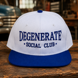 A white and royal blue snapback cap with 'DEGENERATE SOCIAL CLUB' embroidered in blue on the front, resting on a rustic wooden table in a workshop setting.