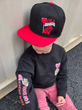 Child wearing a black and red cap, black sweatshirt with pink text, and pink pants on a pavement.